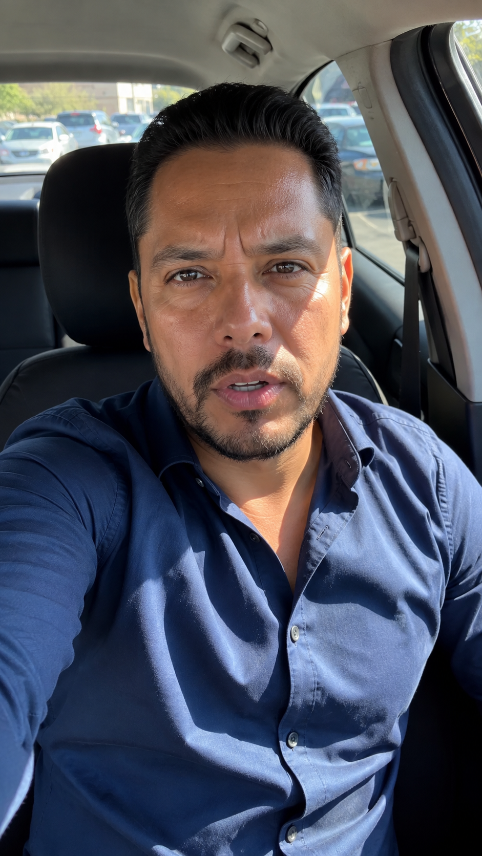 Hombre con camisa azul hablando a cámara dentro de su auto en un estacionamiento